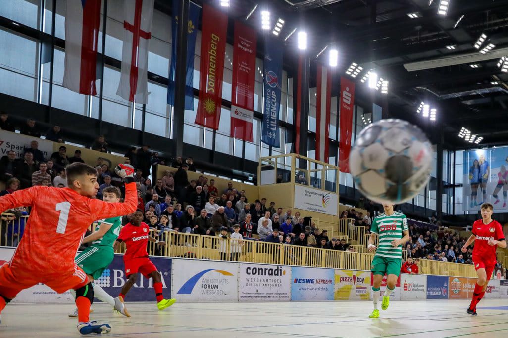 Ein Torwart streckt sich nach dem Ball, dahinter die vollen Ränge der Sporthalle.