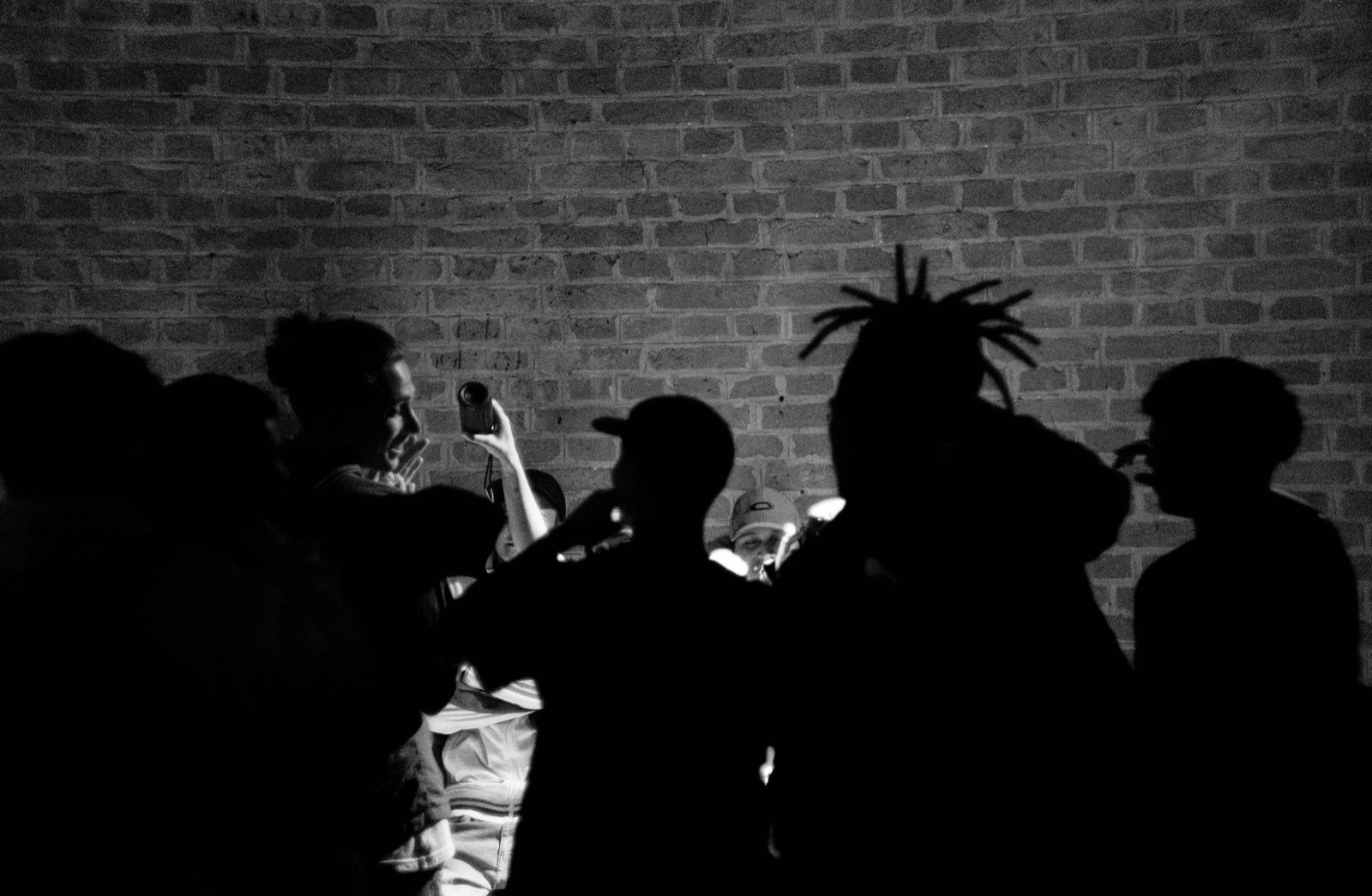 Silhouetten einer Gruppe vor einer Backsteinwand, eine Hand hält ein Mikrofon in die Höhe.
